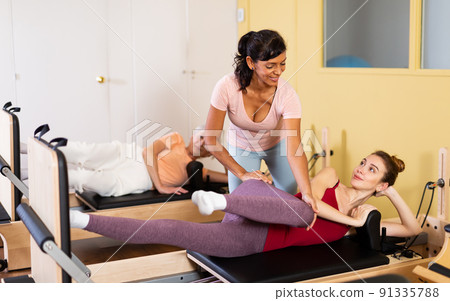 Young woman practicing pilates on reformer with female trainer Young woman practicing pilates on reformer with female trainer 91335788