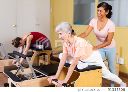 Senior woman practicing pilates on combo chair with Hispanic female trainer Senior woman practicing pilates on combo chair with Hispanic female trainer 91335806