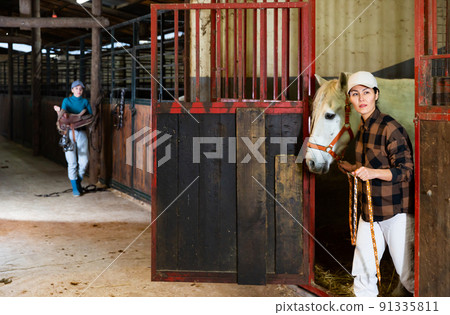 Asian female horse breeder leading white racehorse by bridle out of stall door 91335811