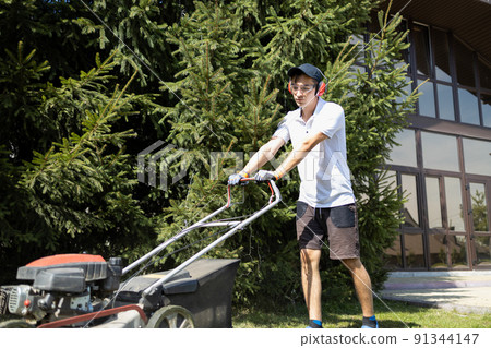 Young man mows a lawn near a large mansion. Worker with a gasoline lawn mower 91344147