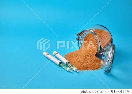 Insulin syringe pens and a glass jar with scattered brown sugar on a blue background with copy space 91344149