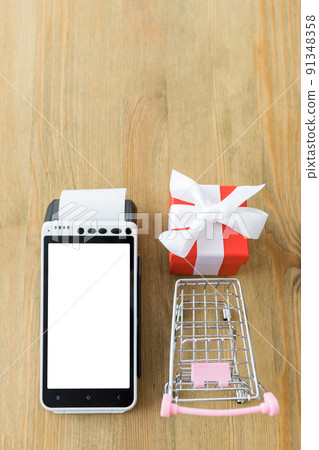 Flat lay of POS terminal with blank screen with miniature shopping cart and red gift box on a wooden table with space for your copy. Valentine's Day shopping concept. Flat lay of POS terminal with blank screen with miniature shopping cart and red gift box on a wooden table with space for your copy. Valentine's Day shopping concept. 91348358