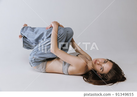 Portrait of young beautiful girl lying on floor, posing in jeans and cotton underwear top isolated over grey studio background. Lifestyle 91349655