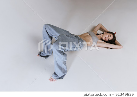 Portrait of young beautiful girl lying on floor, posing in jeans and cotton underwear top isolated over grey studio background. Well-being 91349659