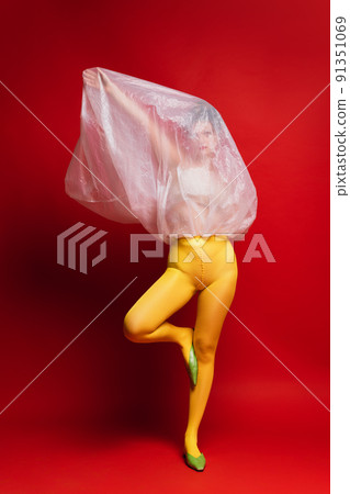 Portrait of young girl in yellow tights with cellophane covering face isolated over red background. Extraordinary art photography. Portrait of young girl in yellow tights with cellophane covering face isolated over red background. Extraordinary art photography. 91351069