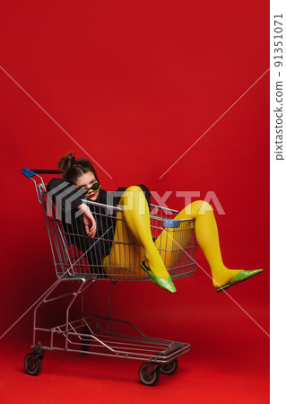 Funny portrait of stylish young girl in yellow tights sitting on shopping trolley, posing isolated over red background Funny portrait of stylish young girl in yellow tights sitting on shopping trolley, posing isolated over red background 91351071