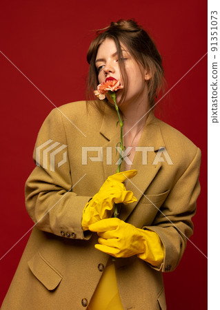 Stylish portrait of young girl with messy hair posing in oversized jacket yellow rubber gloves and flower isolated over red background 91351073