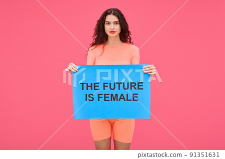 Portrait of young woman holding The future is female poster and looking at camera expressing her rights against pink background Portrait of young woman holding The future is female poster and looking at camera expressing her rights against pink background 91351631