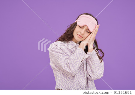 Portrait of young woman in sleep mask closed her eyes and putting head on her hands during dreaming against purple background 91351691