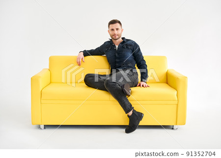 Young trendy man posing sitting on yellow sofa looking to camera 91352704