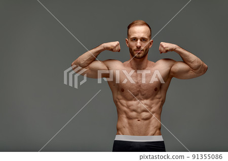 Portrait of a handsome young guy model with hairstyle in a trendy white tank top mock up with a muscular healthy body showing off his biceps muscles on a gray background 91355086