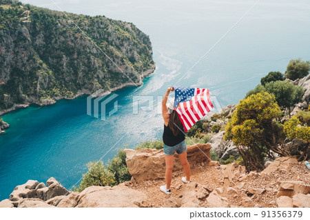 Young woman standing on a rock cliff and waving the US flag while looking at sea beneath. Girl traveller waving the American flag while standing on a mountain top. 4 fourth July Independence Day. 91356379