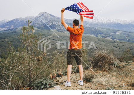 Young man standing on a rock cliff and waving the US flag while looking at mountains in the background. Male traveller waving American flag standing on mountain top. 4 fourth July Independence Day 91356381