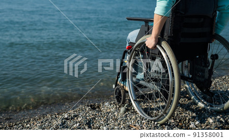 Caucasian woman in a wheelchair on the seashore. Close-up of female hands. Caucasian woman in a wheelchair on the seashore. Close-up of female hands. 91358001