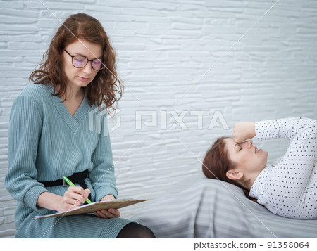 A caucasian woman lies on a couch and expresses her feelings, while a psychologist makes notes on a tablet. A caucasian woman lies on a couch and expresses her feelings, while a psychologist makes notes on a tablet. 91358064