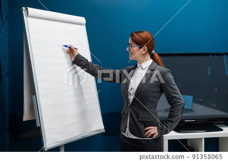 Friendly woman in a suit writes on a blank white board with a marker. Red-haired girl makes a presentation in the office. Beautiful female business coach at a conference. 91358065