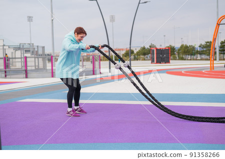 Caucasian woman in a mint sweatshirt is training with battle ropes at the sports ground.  91358266