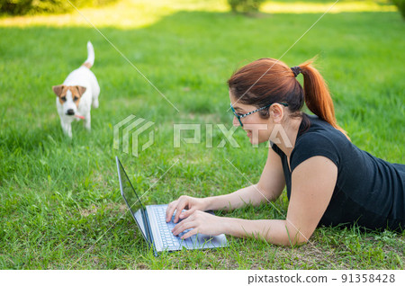 A woman with glasses lies on the green grass in the park with her own dog. A girl blogger maintains a social distance and maintains her blog outdoors. The student is studying remotely on a laptop. 91358428