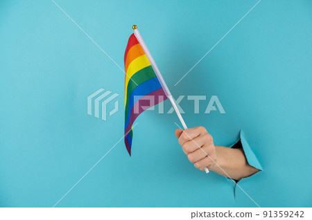 A woman's hand sticks out of a hole in a blue paper background and holds an lgbt flag. A woman's hand sticks out of a hole in a blue paper background and holds an lgbt flag. 91359242