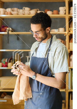 Young multi-ethnic craftsman in apron wiping his hands with towel 91359874
