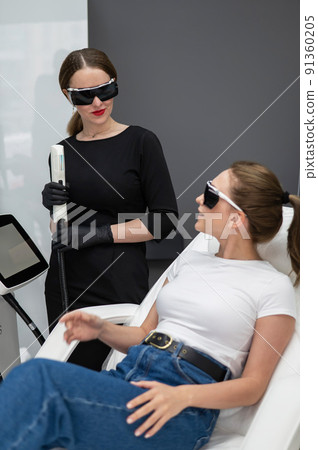 A woman on a couch in goggles undergoing a photorejuvenation procedure. Cosmetologist with a patient. A woman on a couch in goggles undergoing a photorejuvenation procedure. Cosmetologist with a patient. 91360205