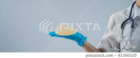 Female doctor aesthetic surgeon holding a breast implant on a white background.  91360320