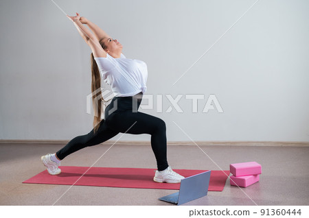 Young Plus Size Woman Stretching At Home Online. Flexible girl practices yoga and watches an online course on a laptop 91360444