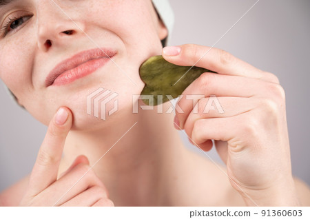 Close-up portrait of a young woman massaging her face with a gouache scraper.  91360603