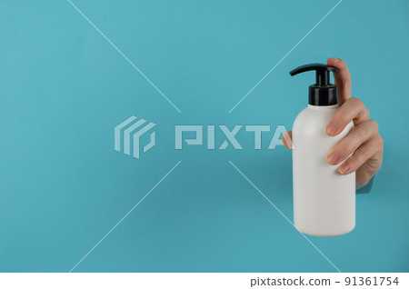 A woman's hand sticks out of a hole in a blue paper background and holds a skin lotion pump. A woman's hand sticks out of a hole in a blue paper background and holds a skin lotion pump. 91361754