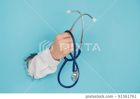 A woman's hand sticking out of a hole on a blue paper background holds a stethophonendoscope. 91361761