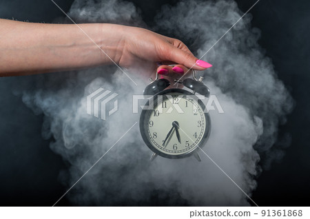 A woman holds an alarm clock in a studio full of smoke. White fog enveloped a round retro mechanical watch. A woman holds an alarm clock in a studio full of smoke. White fog enveloped a round retro mechanical watch. 91361868