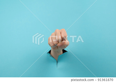 A woman's hand sticking out of a hole from a blue background shows a fist. A woman's hand sticking out of a hole from a blue background shows a fist. 91361870