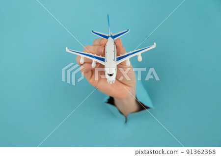 A female hand sticking out of a hole from a blue background holds a model of an airplane. A female hand sticking out of a hole from a blue background holds a model of an airplane. 91362368