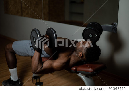 Shirtless afro american man doing exercises with dumbbells lying on bench in gym.  91362511