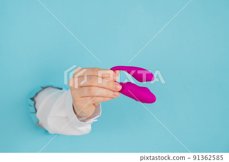 A woman's hand holds a pink sex toy through a hole in a paper blue background. A woman's hand holds a pink sex toy through a hole in a paper blue background. 91362585