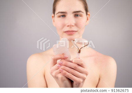 Caucasian woman holding pink roller massager and gouache scraper on white background.  91362675
