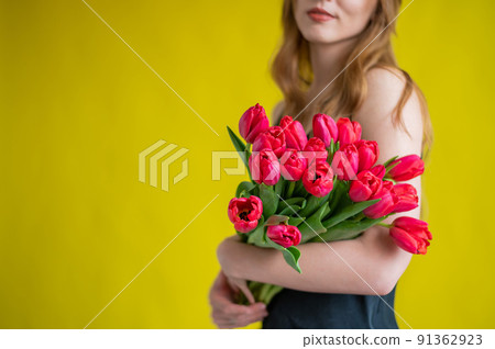 Unrecognizable woman with a bouquet of red tulips on a yellow background. A girl in a black dress holds an armful of flowers. A gift for International Women's Day. Spring holiday. Unrecognizable woman with a bouquet of red tulips on a yellow background. A girl in a black dress holds an armful of flowers. A gift for International Women's Day. Spring holiday. 91362923