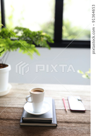 White coffee cup and phone with notebooks and plant pot on brown wooden table in front of window White coffee cup and phone with notebooks and plant pot on brown wooden table in front of window 91364653