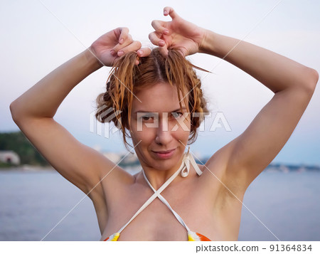 A funny girl in a bikini is holding her wet pigtails. Goofing around on a summer beach weekend. 91364834