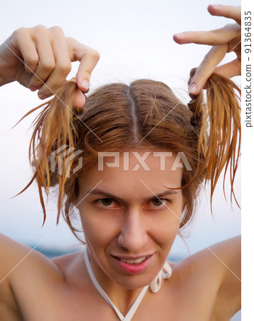 A funny girl with a stern look holds her wet pigtails. Summer sea holidays. 91364835