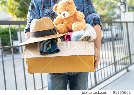 Holding clothing donation box with used clothes and doll at home to support help for poor people in the world. Holding clothing donation box with used clothes and doll at home to support help for poor people in the world. 91365601