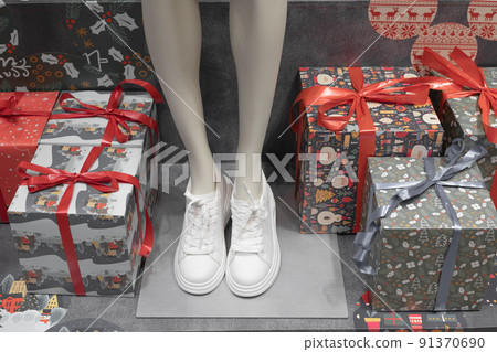 Female leg mannequin in white sneakers on display in the middle of gift boxes. Christmas shop decor. Holiday shopping. Female leg mannequin in white sneakers on display in the middle of gift boxes. Christmas shop decor. Holiday shopping. 91370690