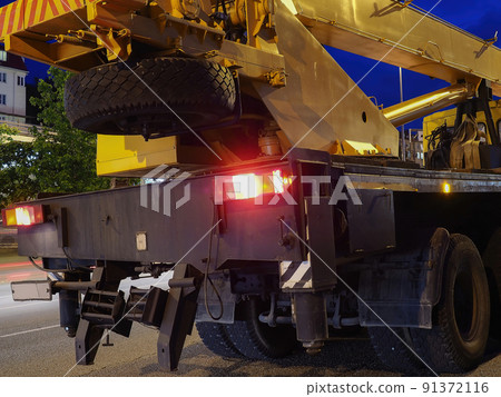 A yellow-black truck crane with headlights on stands on a night road. Road works A yellow-black truck crane with headlights on stands on a night road. Road works 91372116
