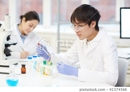 Serious young Asian lab technician in protective gloves and white coat sitting at table in laboratory and using pipette while adding reagent into test tube Serious young Asian lab technician in protective gloves and white coat sitting at table in laboratory and using pipette while adding reagent into test tube 91374566
