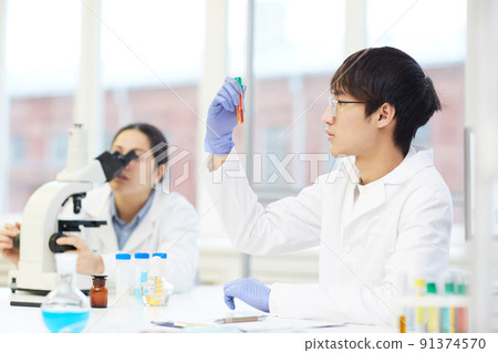Serious young Asian lab employee in white coat sitting at table with laboratory tools and looking at liquid in test tube 91374570