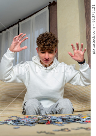 Young caucasian man with curly hair sitting on the sofa at home with his arms raised in protest at the difficulty of assembling a puzzle of 1000 pieces 91375121