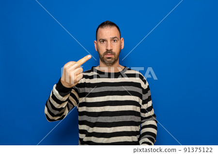 Hispanic man with beard wearing striped sweater standing over isolated blue background Showing middle finger, impolite and rude fuck expression. 91375122