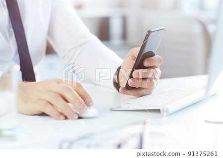Close-up of unrecognizable businessman in formal shirt sitting at desk and checking smartphone while working in office Close-up of unrecognizable businessman in formal shirt sitting at desk and checking smartphone while working in office 91375261