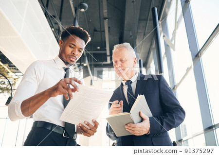 Below view of serious experienced manager in suit holding book and discussing project plan with intern in office Below view of serious experienced manager in suit holding book and discussing project plan with intern in office 91375297