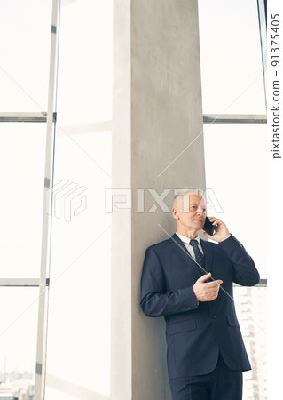 Content handsome mature businessman in formal suit leaning on column in lobby while talking by cellphone Content handsome mature businessman in formal suit leaning on column in lobby while talking by cellphone 91375405